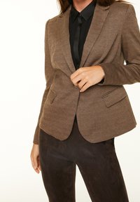 Hnědý kostkovaný blazer s klopami, dvěma předními kapsami a přiléhavým střihem, kombinovaný s černou košilí na knoflíky a tmavými kalhotami.