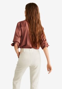 Bruine satijnen blouse met opgerolde mouwen, gedragen met witte high-waisted broeken. Lang bruin haar valt over de rug. Minimalistische setting.