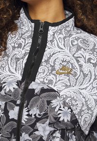 Nike-jacka med ett svart och vitt blommigt spetsmönster, dragkedja, med guldlogotyp och textilier med struktur.