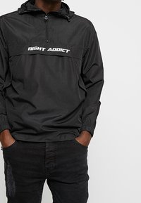 Zwarte windjack met een ritssluiting bij de kraag, elastische manchetten en een wit "NIGHT ADDICT" logo op de borst. Gladde textuur, lichtgewicht materiaal.