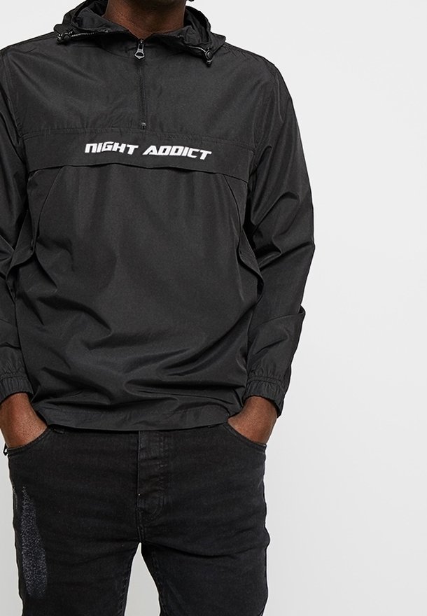 Zwarte windjack met een ritssluiting bij de kraag, elastische manchetten en een wit "NIGHT ADDICT" logo op de borst. Gladde textuur, lichtgewicht materiaal.