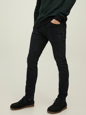 Dunne zwarte jeans, katoenen stof, lichte stretch, voorzakken, gedragen met donkere suède schoenen en een groene gebreide trui met zwarte mouwen.