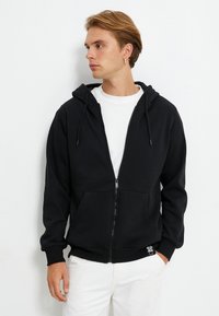 Sudadera con cremallera negra, con cordones, bolsillos delanteros y puños acanalados. Llevada sobre una camiseta blanca; textura suave y diseño casual.