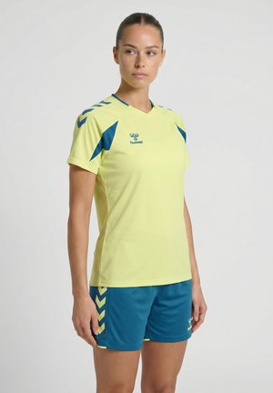 Kobieta w żółto-niebieskiej sportowej koszulce i pasujących szortach, stojąca z rozluźnionymi ramionami, lekko patrząca na bok na jednolitym tle.