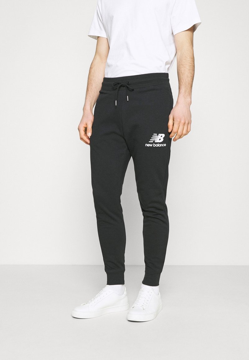 Zwarte katoenen joggers met een aansluitend ontwerp, voorzien van een wit "New Balance"-logo, trekkoord in de taille en geribbelde boorden. Draagt witte sneakers.
