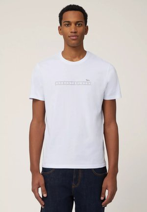 Giovane uomo in piedi, indossa una maglietta bianca a maniche corte con la scritta "Harmont & Blaine", abbinata a jeans blu scuro.