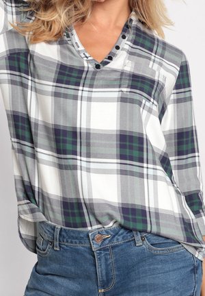 Femme portant une chemise à carreaux verte, marine et blanche à manches longues avec des boutons au niveau du col, assortie avec un jean en denim bleu.