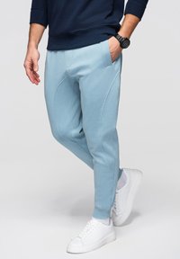 Mand iført lyseblå joggebukser, hvide sneakers, marineblå sweatshirt og sort armbåndsur, stående med den ene hånd i lommen.