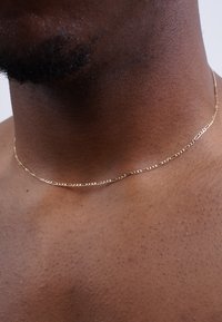 Goldene Kettenhalskette mit verlängerten Gliedern und strukturierten Oberflächen, die auf einem nackten Hals ruhen, vor einem neutralen Hintergrund.
