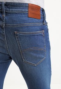 Mörkblå denimjeans har en brun läderlogg, två bakfickor med böjd sömnad och en något bleknad struktur.