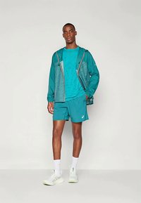 Teal urheiluasu, johon kuuluu kevyt huppari, lyhythihainen paita ja shortsit, joita täydentävät valkoiset lenkkarit ja sukat.