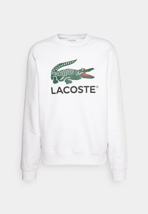 Balta medvilninė džemperis su žaliu ir raudonu krokodilo logotipu ir užrašytu "LACOSTE" žemiau, su raukiniais rankogaliuose ir apačioje.