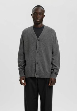 Mand iført en tekstureret grå knappet cardigan over en sort skjorte og løse sorte bukser, stående mod en ensartet lys baggrund.