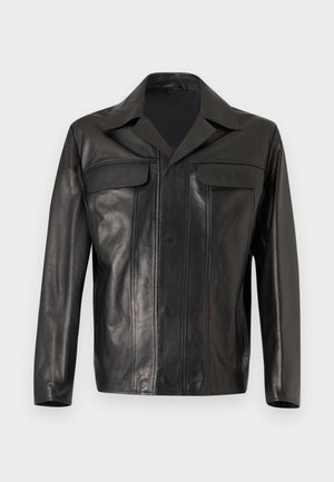 Man met kort blond haar die een zwarte leren jas draagt over een grijze blazer en wit overhemd, zwarte wijde broek en lichtbruine schoenen.
