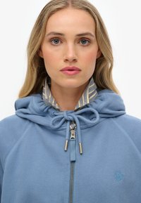 Blauwe zip-up hoodie met een capuchon met trekkoord, voorzien van een contrasterende gestreepte binnenvoering en een geborduurd logo op de borst.