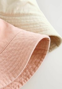 Två bucket hats: en ljus beige och en mjuk rosa. Båda har ett texturerat tyg med ett sytt mönster längs brätten.