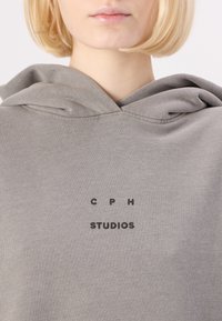 Hanorac gri dintr-un material moale, având o mare buzunar frontal și textul "C P H" deasupra "STUDIOS" brodat cu fir negru.