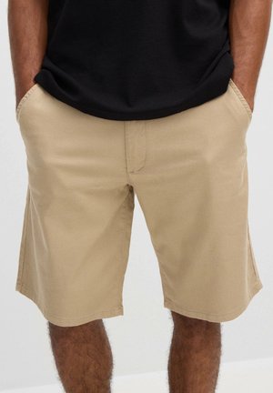 Hombre con pantalones cortos beige hasta la rodilla y una camisa negra, con las manos en los bolsillos, de pie frente a un fondo claro y liso.