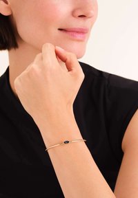 Pulsera de oro con un diseño liso y minimalista que presenta un único acento de gema azul. Lleva puesta en una muñeca sobre un fondo neutro.