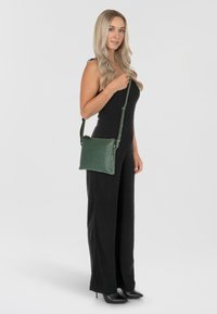 Borsa a tracolla verde in pelle con superficie texturizzata, chiusura zip e tracolla regolabile. Indossata da un modello che indossa una tuta nera.