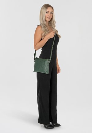 Borsa a tracolla verde in pelle con superficie texturizzata, chiusura zip e tracolla regolabile. Indossata da un modello che indossa una tuta nera.