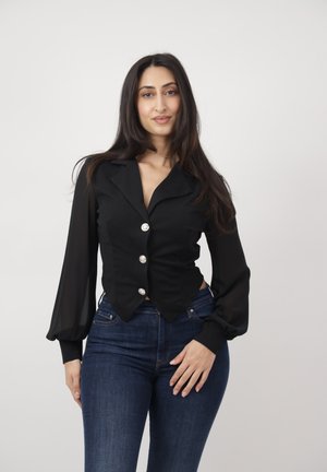 Mujer con cabello largo y oscuro, vistiendo una blusa negra abotonada con mangas transparentes y jeans azul oscuro, de pie contra un fondo claro y liso.