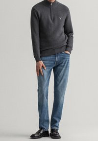 Pull à col zippé gris foncé avec un motif texturé, associé à un jean bleu et des chaussures en cuir noires. Le pull possède des poignets et un ourlet côtelés.
