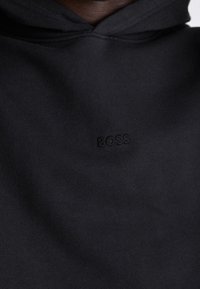 Svart hoodie med en liten "BOSS"-logotyp broderad på bröstet, som visar överkroppen och halslinningen.