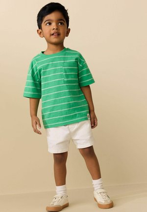 Ung dreng iført en grønstribet t-shirt, hvide shorts, hvide sokker og beige sneakers stående mod en beige baggrund.