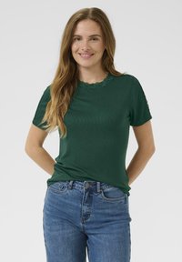 T-shirt vert à côtes avec un col rond bordé de dentelle et des manches courtes, associé à un jean bleu. Texture lisse, coupe décontractée.