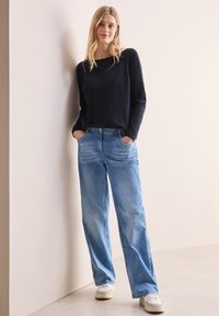 Dunkelblauer Pullover mit langen Ärmeln und Rundhalsausschnitt, kombiniert mit hellblauen geraden Jeans und weißen Sneakers, steht vor einem neutralen Hintergrund.