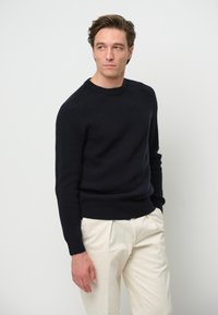 Marinblå ribbad stickad tröja med rund hals och långa ärmar, kombinerad med ljusa, högmidjade byxor, mot en neutral bakgrund.