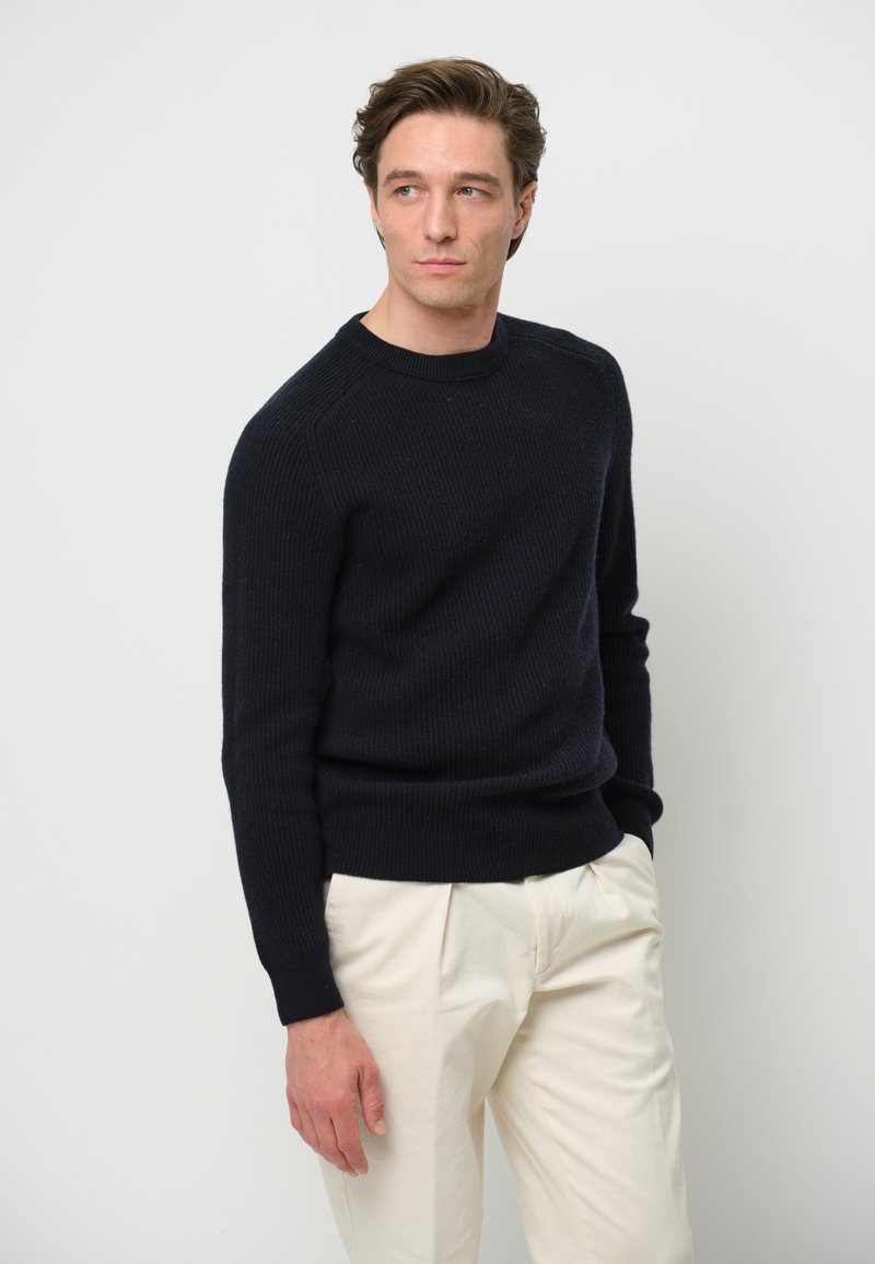 Marinblå ribbad stickad tröja med rund hals och långa ärmar, kombinerad med ljusa, högmidjade byxor, mot en neutral bakgrund.