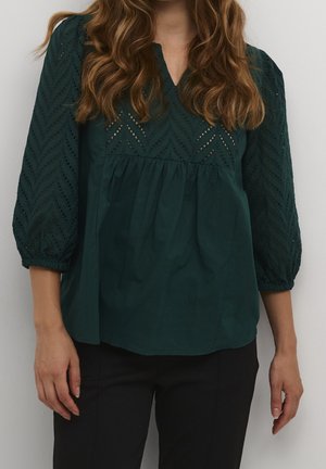 Vrouw draagt een donkergroene blouse met mouwen met oogjespatroon en een V-hals, gecombineerd met een zwarte broek, staande tegen een effen achtergrond.