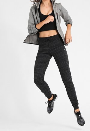 Woman jumping in black sports bra, gray zip-up jacket, dark gray joggers, and black athletic shoes against white background.