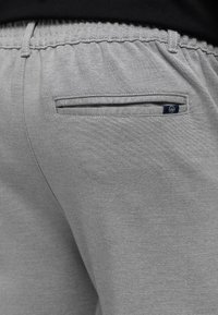 Harmaat kangashousut, joissa on joustava vyötärönauha ja vaakasuora takatasku, jossa on pieni mustavalkoinen logolappu.