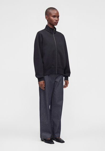 Personne portant une veste noire zippée, un pantalon large foncé et des chaussures plates noires, debout devant un fond clair uni.