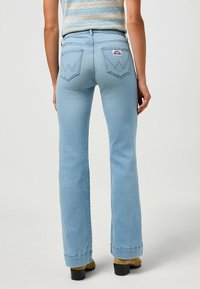 Mulher vestindo jeans bootcut azuis claros e uma parte de cima sem mangas, de pé com as costas viradas para a câmara contra um fundo simples.