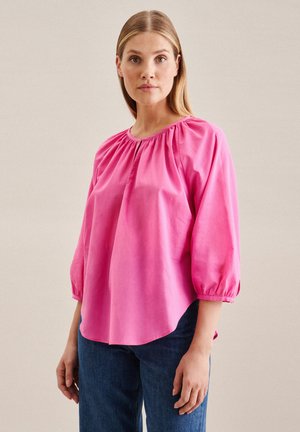 Rosa, leichte Bluse mit rundem Ausschnitt, gerafften Details an den Schultern und dreiviertellangen Puffärmeln. Getragen mit dunklen Jeans.