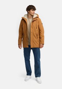 Brūns virsjakas apvalks ar pūkainu cepures oderi, ar rāvējslēdzēju un sānu kabatu. Valkāts pāri bēšam augšdaļai, kopā ar ziliem džinsiem un baltiem sporta apaviem.