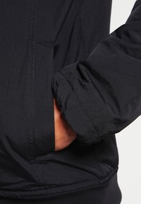 Giacca nera leggera con un tessuto texture, dotata di una tasca sul petto e polsini elasticizzati, che enfatizza praticità e comfort.