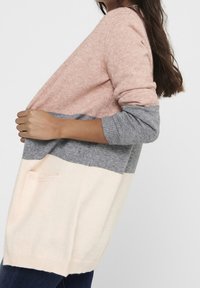 Femme portant un long cardigan en blocs de couleurs rose, gris et crème, tenant les bords avant, avec des cheveux bruns sur une épaule.