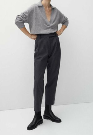 Person trägt einen grauen Pullover mit offenem Kragen, dunkel-hochgeschnittene Hose und schwarze Stiefeletten, steht mit den Händen in der Hüfte vor einfachem Hintergrund.