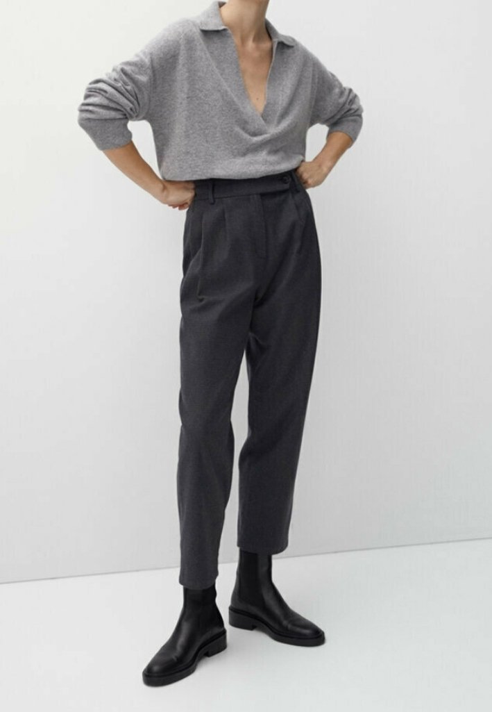 Personne portant un pull gris à col ouvert, un pantalon foncé taille haute et des bottines noires, debout les mains sur les hanches devant un fond uni.