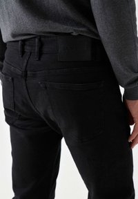 Crne traperice od denima s teksturiranim završetkom, s kožnom etiketu na pojasu i dva stražnja džepa.