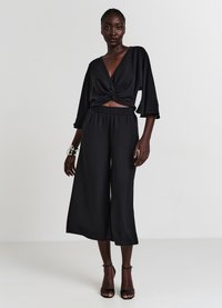 Blusa corta nera con dettaglio a nodo in vita, pantaloni neri a gamba larga con tessuto liscio, abbinati a sandali neri con tacco.