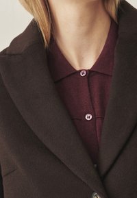 Brauner Wollmantel mit breitem Reverskragen über einem burgunderrot gestrickten Shirt, mit kontrastierenden lila Knöpfen. Glatte Textur und maßgeschneiderte Passform.