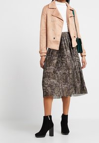 Tan farvet faux suede jakke med sølvknapper, hvid top, mønstret plissee-nederdel i mørke nuancer og sorte ankelstøvler med blokhæl.