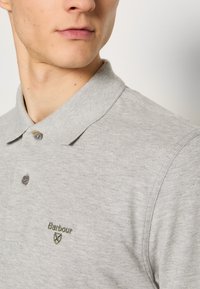 Γκρι πουκάμισο πόλο με κλασικό γιακά, δύο κουμπιά και πράσινο κεντημένο λογότυπο στο αριστερό στήθος. Μαλακό ύφασμα με υφή.