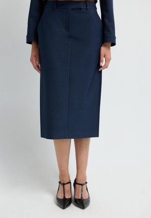 Donna che indossa una gonna a tubino blu navy lunga fino al ginocchio con una maglietta coordinata e décolleté neri in vernice con cinturino a T, in piedi contro un muro semplice.
